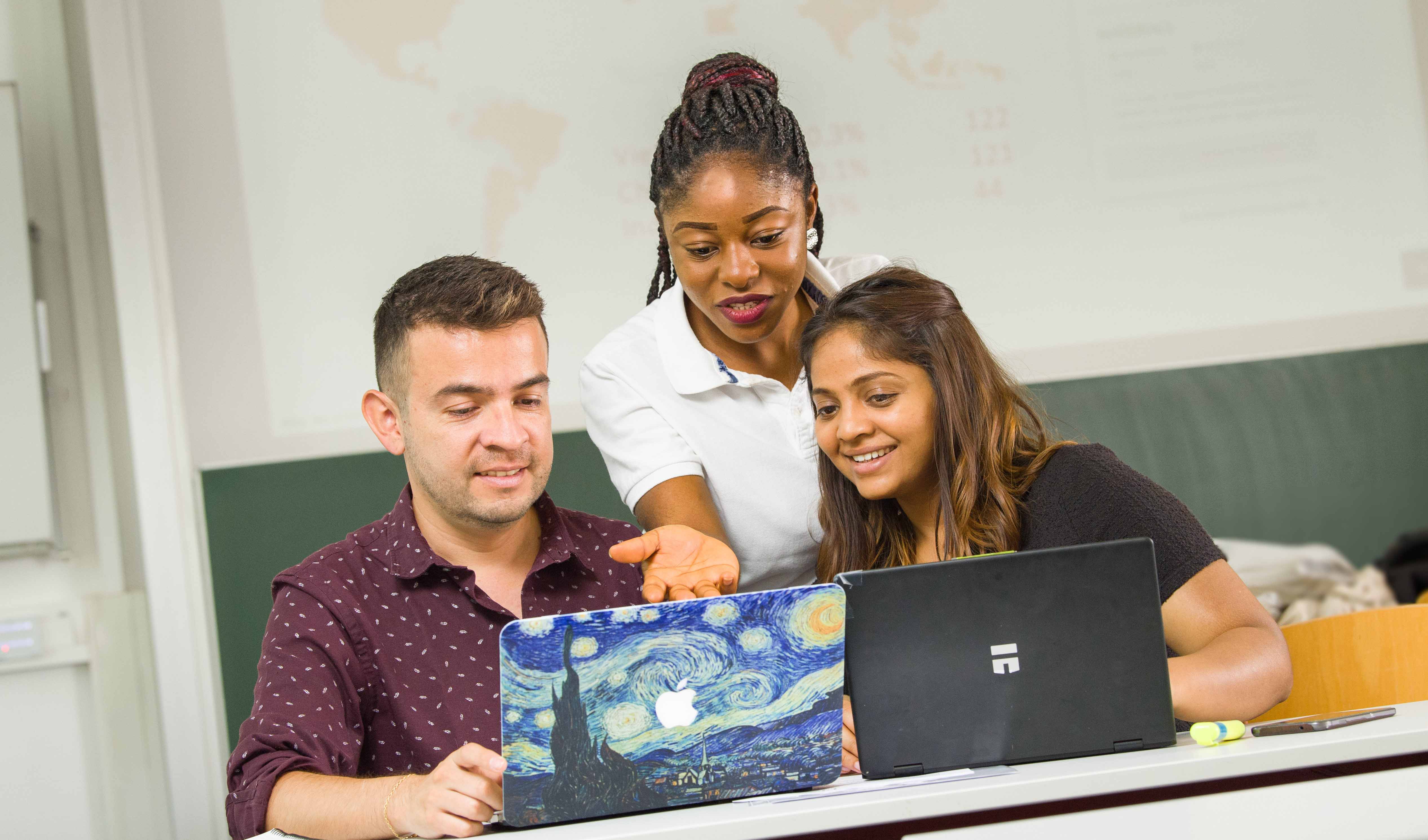 3 internationale Studierende des Studiengangs International Management sitzen im Seminarraum und schauen auf 2 Laptops.