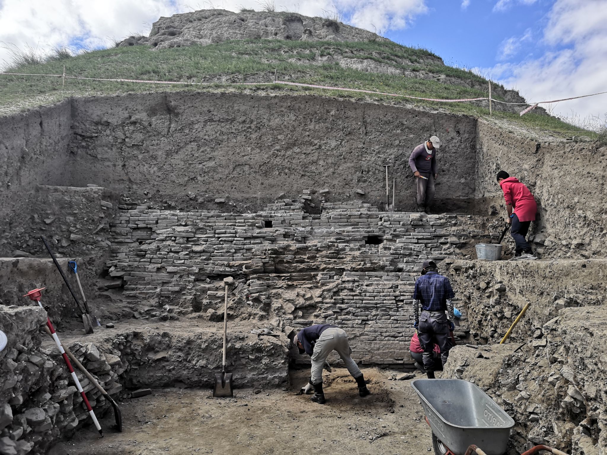 Das Bild zeigt eine archäologische Ausgrabung. In einem großen Schacht mit senkrechten Wänden ist eine historische Mauer und mehrere, an der Freilegung arbeitende Personen zu sehen.