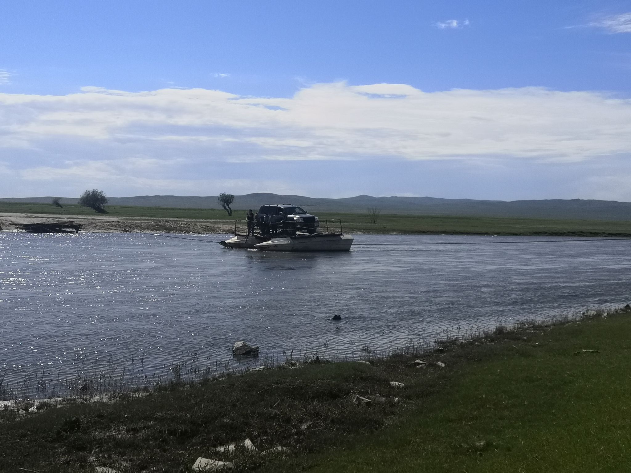 Das Bild zeigt, wie ein Auto auf einer einfachen Fähre aus zwei Pontons an einem Seil über einen Fluss gezogen wird.