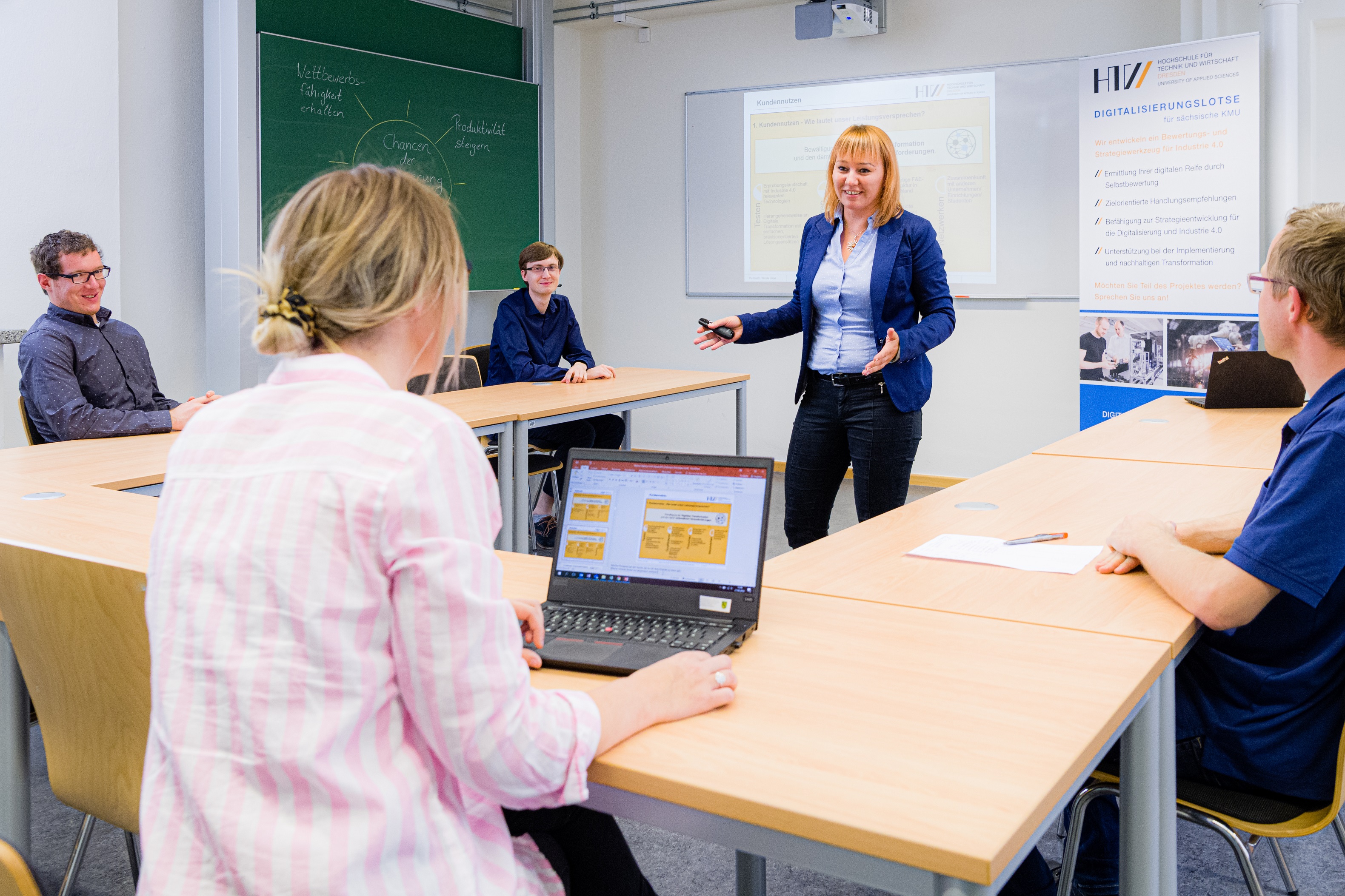 Eine Frau steht bei einem Workshop vor einer Präsentation, mehrere Menschen schauen sitzend zu ihr.