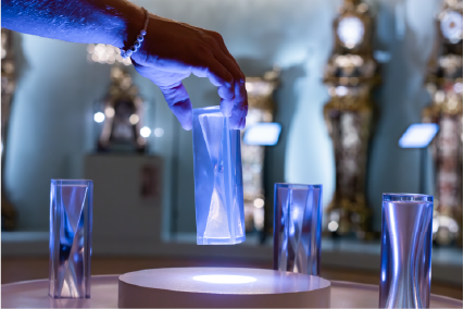 Blick in einen Ausstellungsraum in dem ein Exponat aus mehreren Glaskörpern im Vordergrund stehen und eines mit einer Hand versetzt wird.