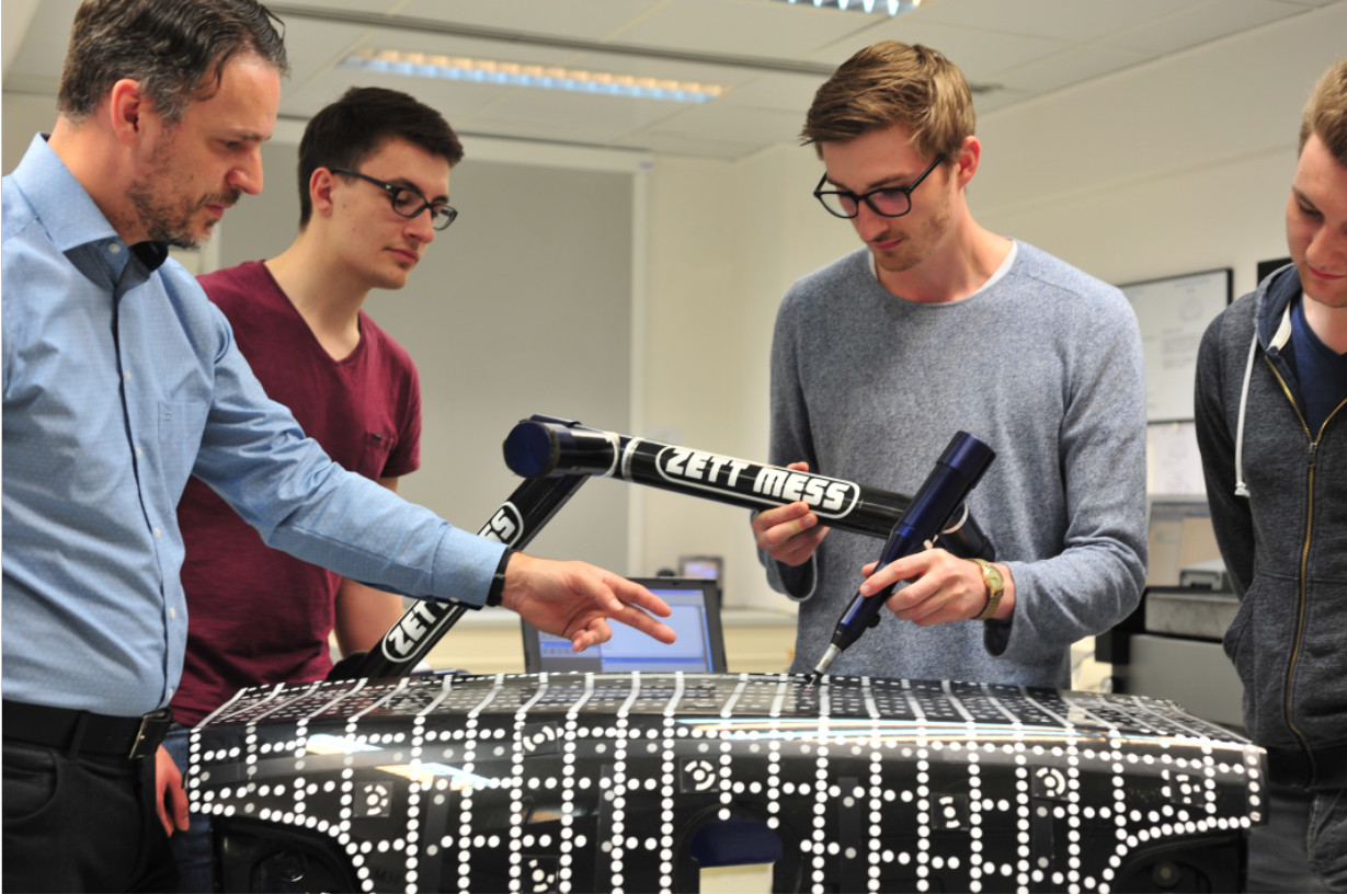 Drei Studierende messen unter Anleitung des Betreuers mit einem Gelenkarmmessgerät die Oberflächengeometrie eines PKW-Kofferraumdeckels.