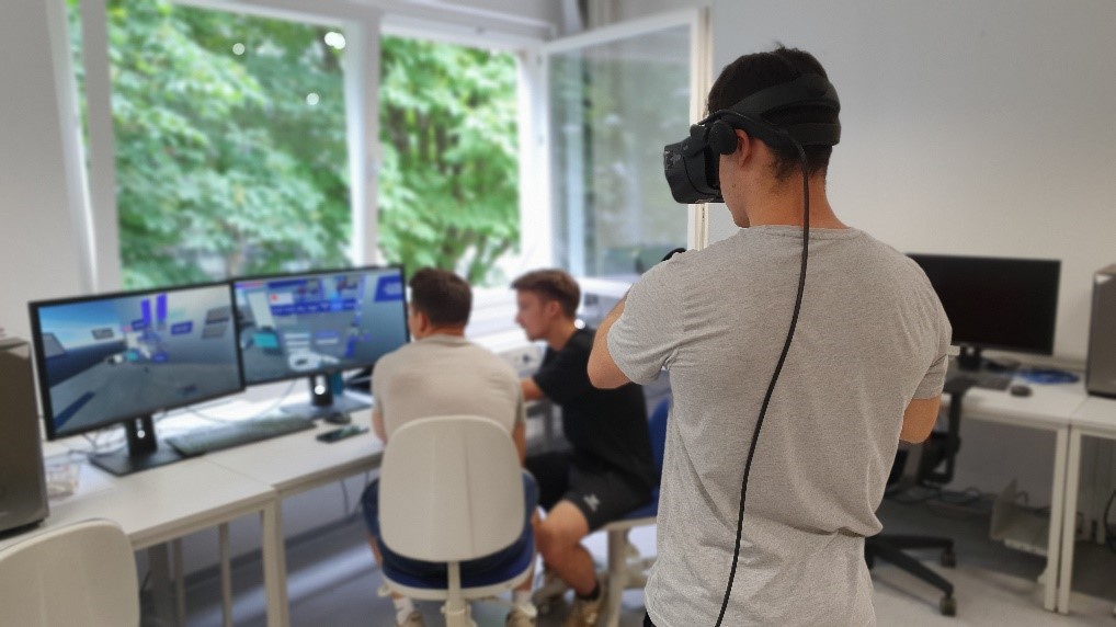 Studierende arbeiten an einem PC-Arbeitsplatz, junger Mann trägt VR-Brille