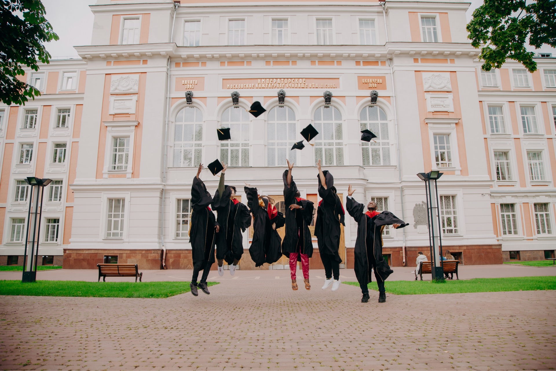 Photo; In the background you can see a white-apricot-colored building; right and left are some branches of trees with green foliage; in the middle of the picture are 6 graduates in black robes. They jump and throw black doctor hats into the air.