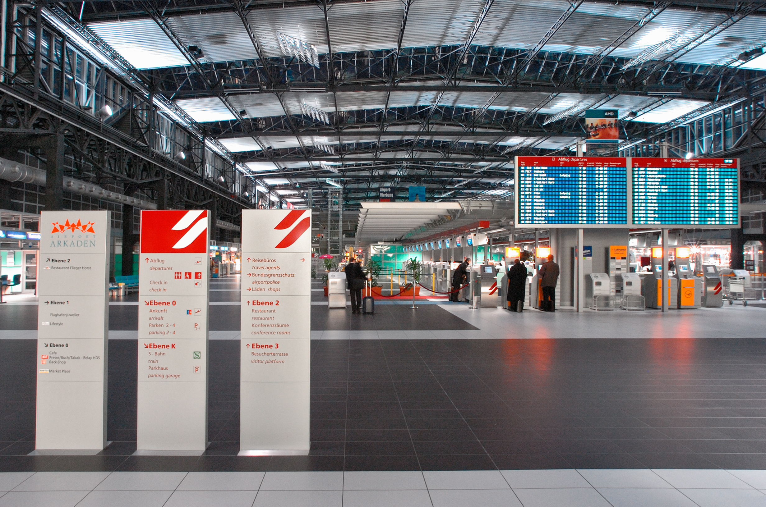 Foto; Am Flughafen; Mittig ist die blaue Anzeigetafel mit den An- und Anflügen zu sehen; rechts sind Schalter zum Einchecken und einige Personen warten davor.