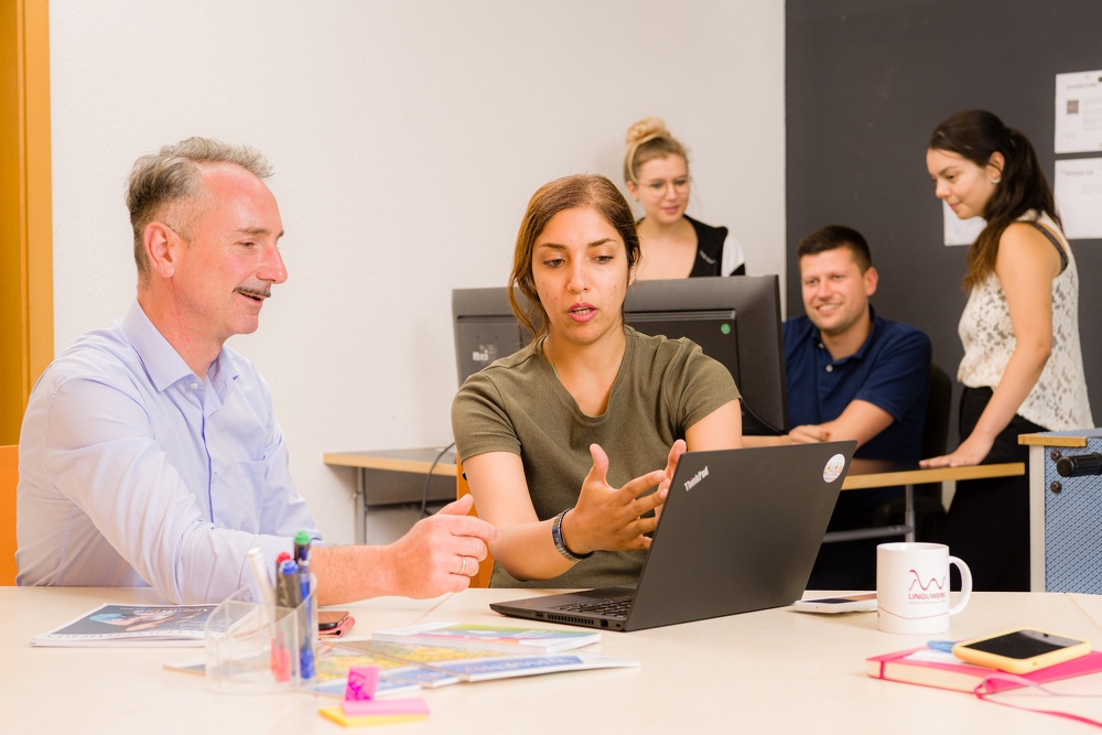 Ein Mann und eine Frau im Vordergrund am Laptop, dahinter 3 Personen sitzen an einem Computer.