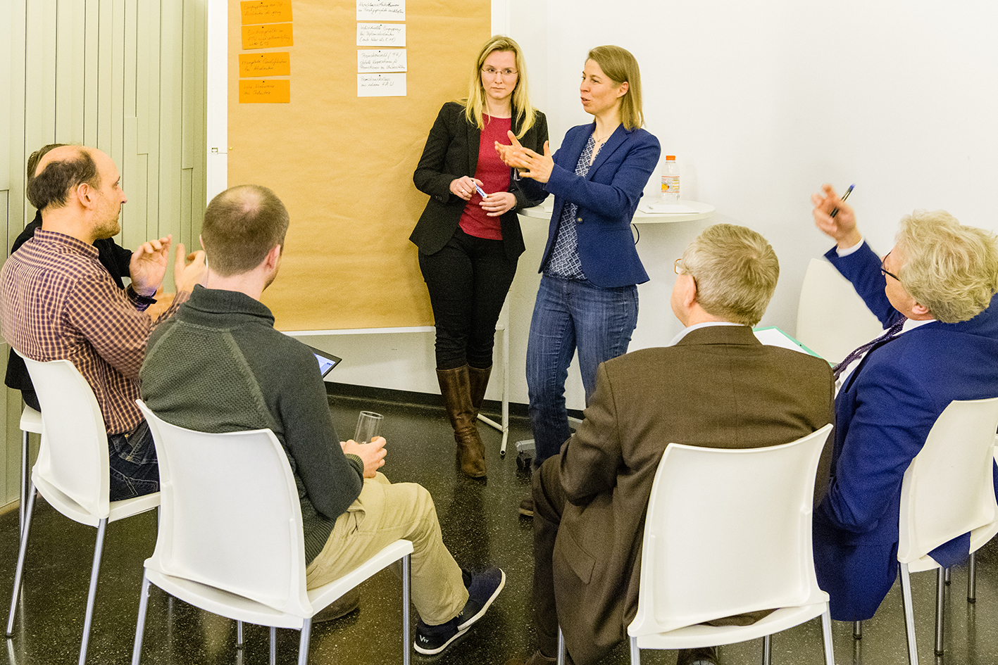 Mehrere Personen sitzen im Halbkreis. Zwei Frauen stehen vor dem Publikum und diskutieren.