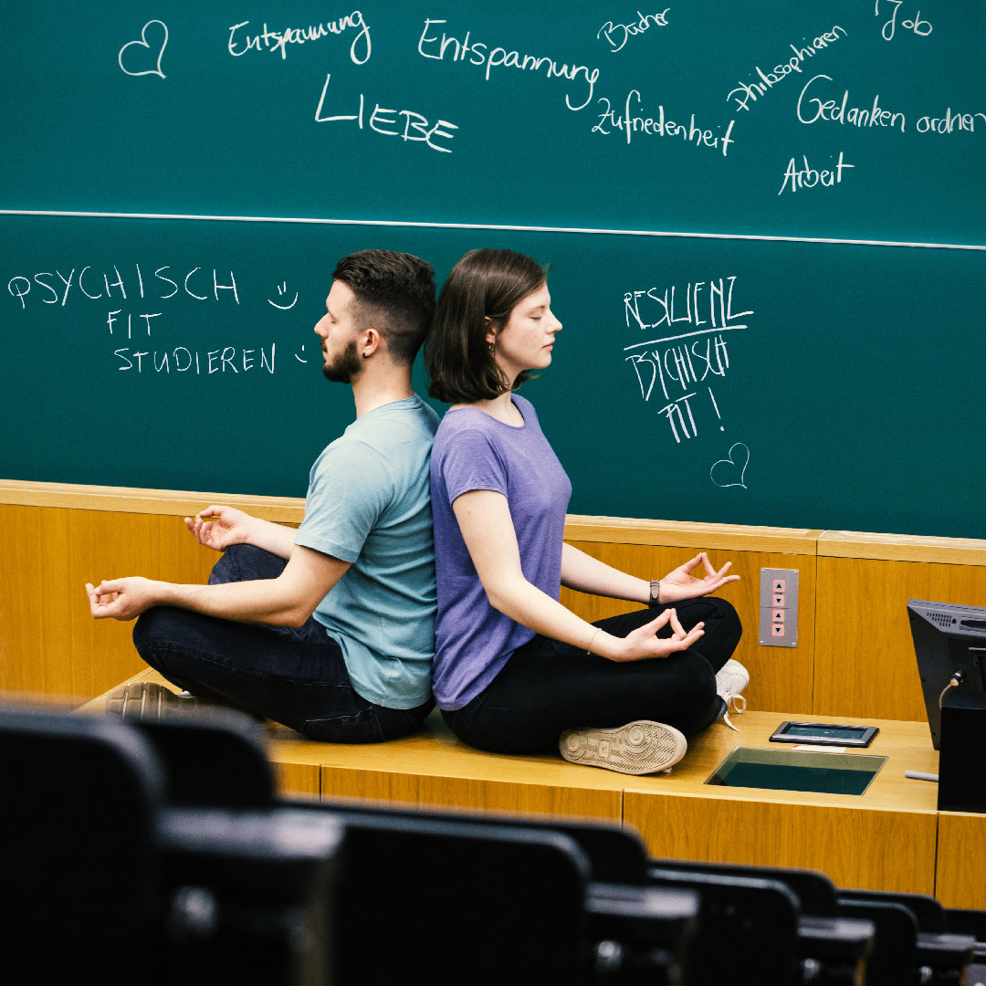 Zwei junge Erwachsene sitzen in einer Meditationshaltung auf dem Präsentationspult in einem Hörsaal