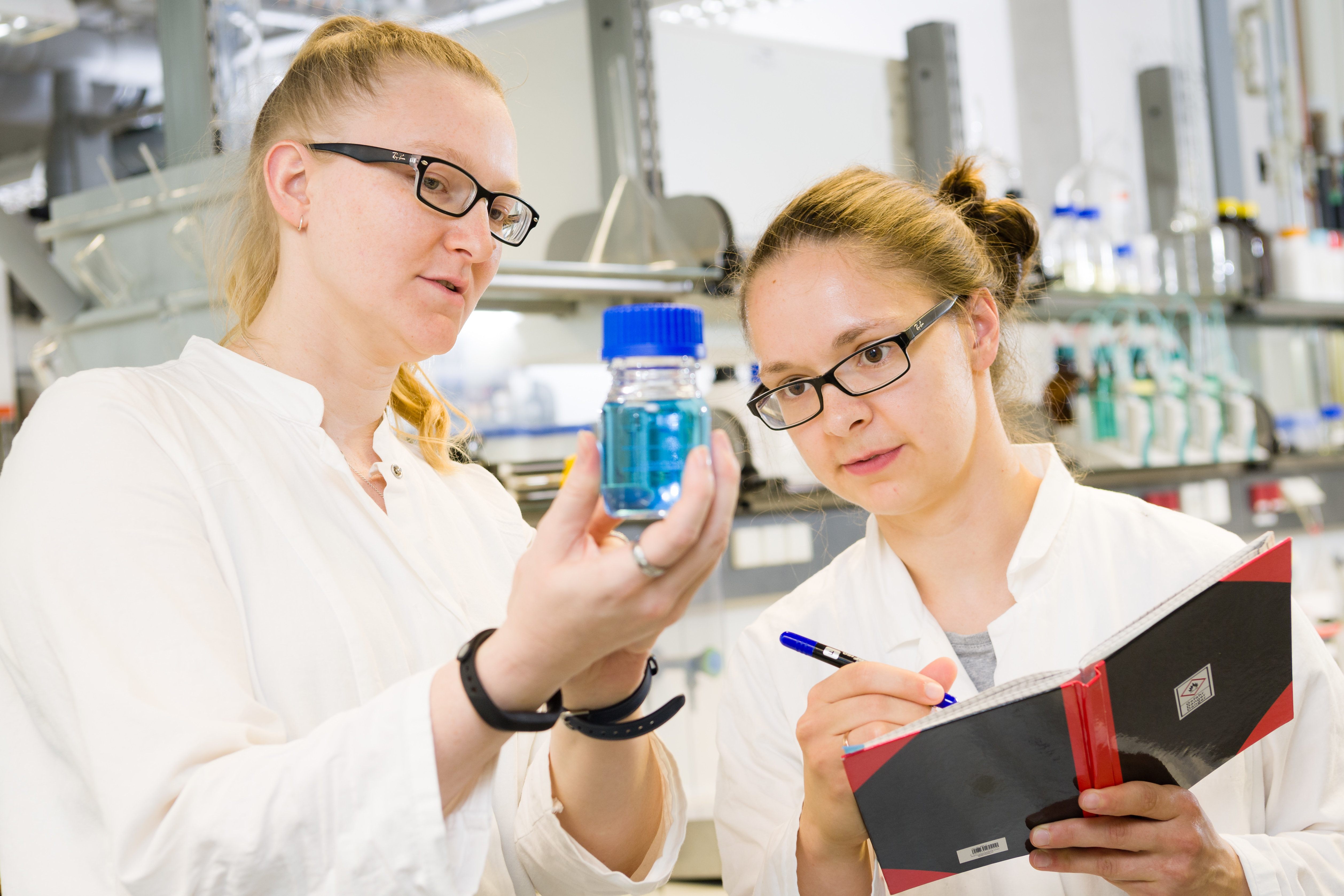 [Translate to English:] 2 Studentinnen schauen sich im Labor gefülltes Fläschchen an