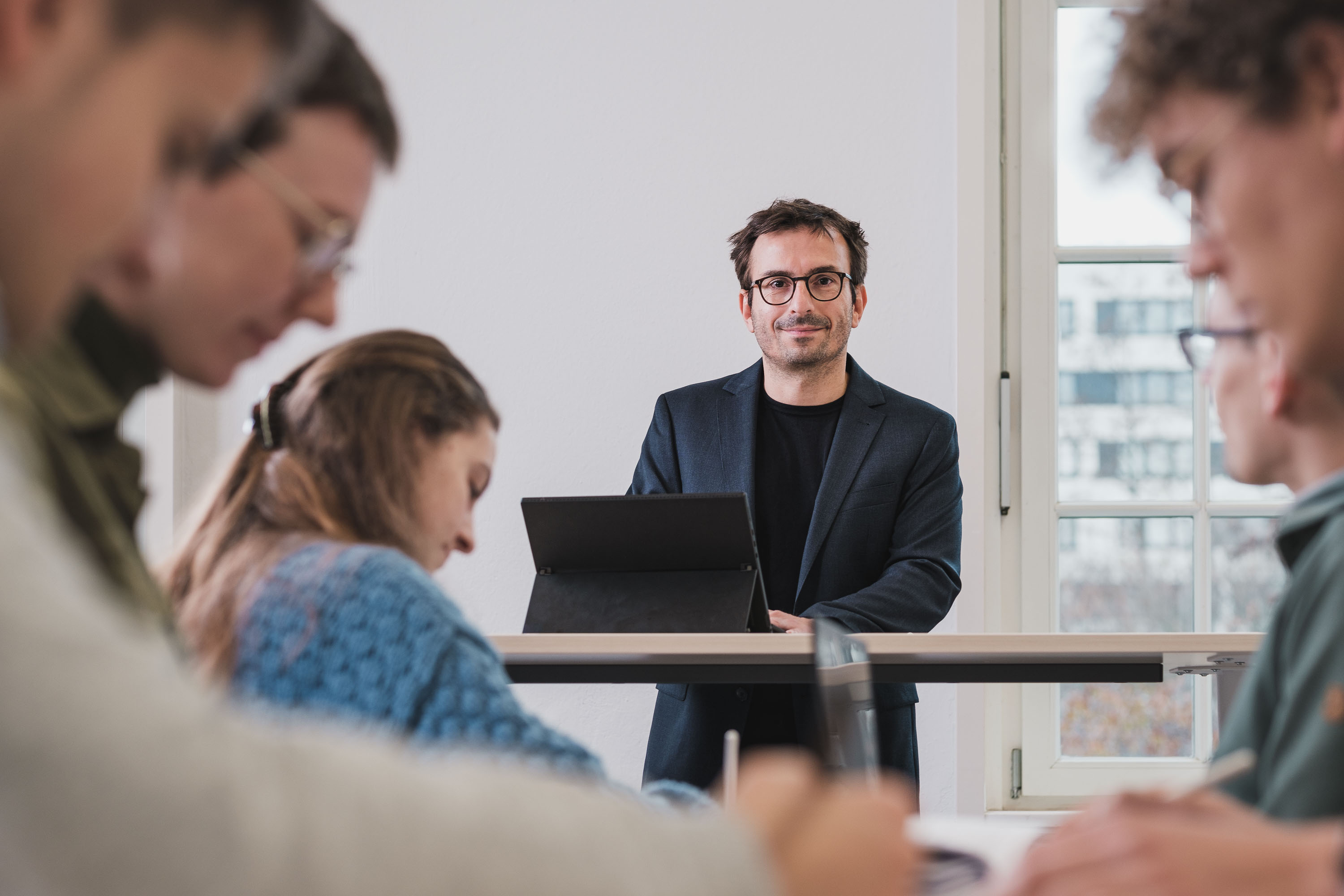 [Translate to English:] Professor blickt direkt in die Kamera umgeben von Studierenden