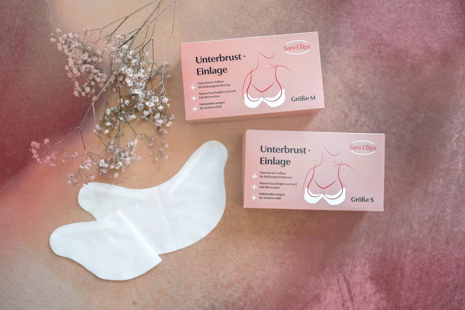 zwei Unterbrustfalteneinlagen liegen auf einem Tisch, daneben zwei geschlossene Packungen von Sani Ellips