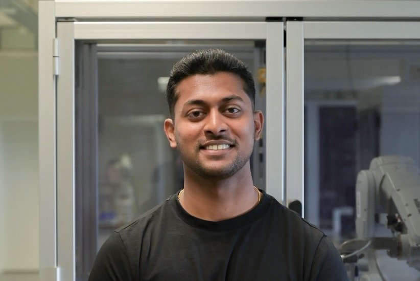 Gunalan Senthil blickt in die KAmera. Im Hintergrund ist eine Roboterzelle zu sehen.