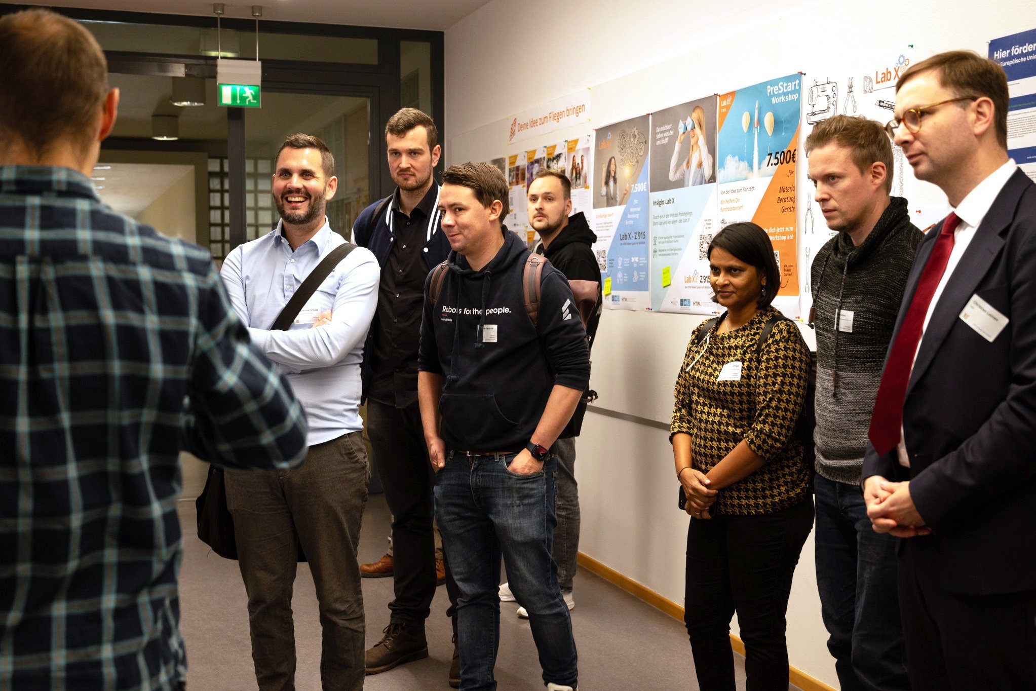 Menschen stehen vor einer Plakatwand und hören interessiert einem Vortragenden zu