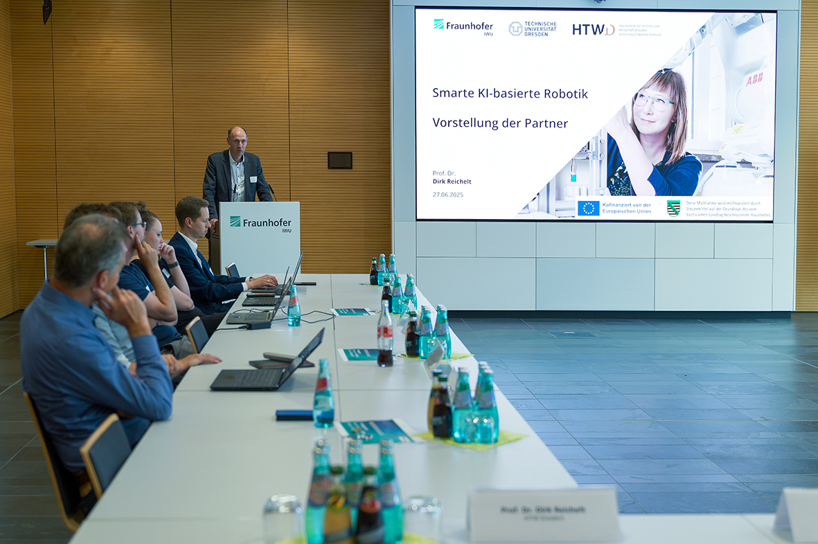 Ein großer MOnitor rechts im Bild, auf dem eine Präsentation angezeigt wird. Links steht Prof. Reichelt am Rednerpult. Im Vordergrund ein Tisch mit Getränken an dem Zuhörende sitzen.