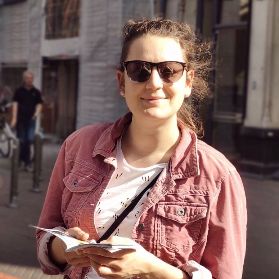 Foto; Studentin Anna aus dem Studiengang Wirtschaftsingenieurwesen. Anna hat rauche Haare, die zusammengebunden sind, trägt eine Sonnenbrille, eine rote Jacke, darunter ein weißes T-Shirt mit schwarzem Muster. SIe hat eine Tasche umgehangen und hält einen Zettel in der Hand, dabei lächelt sie. Im Hintergrund ist ein historisches Gebäude zu sehen.