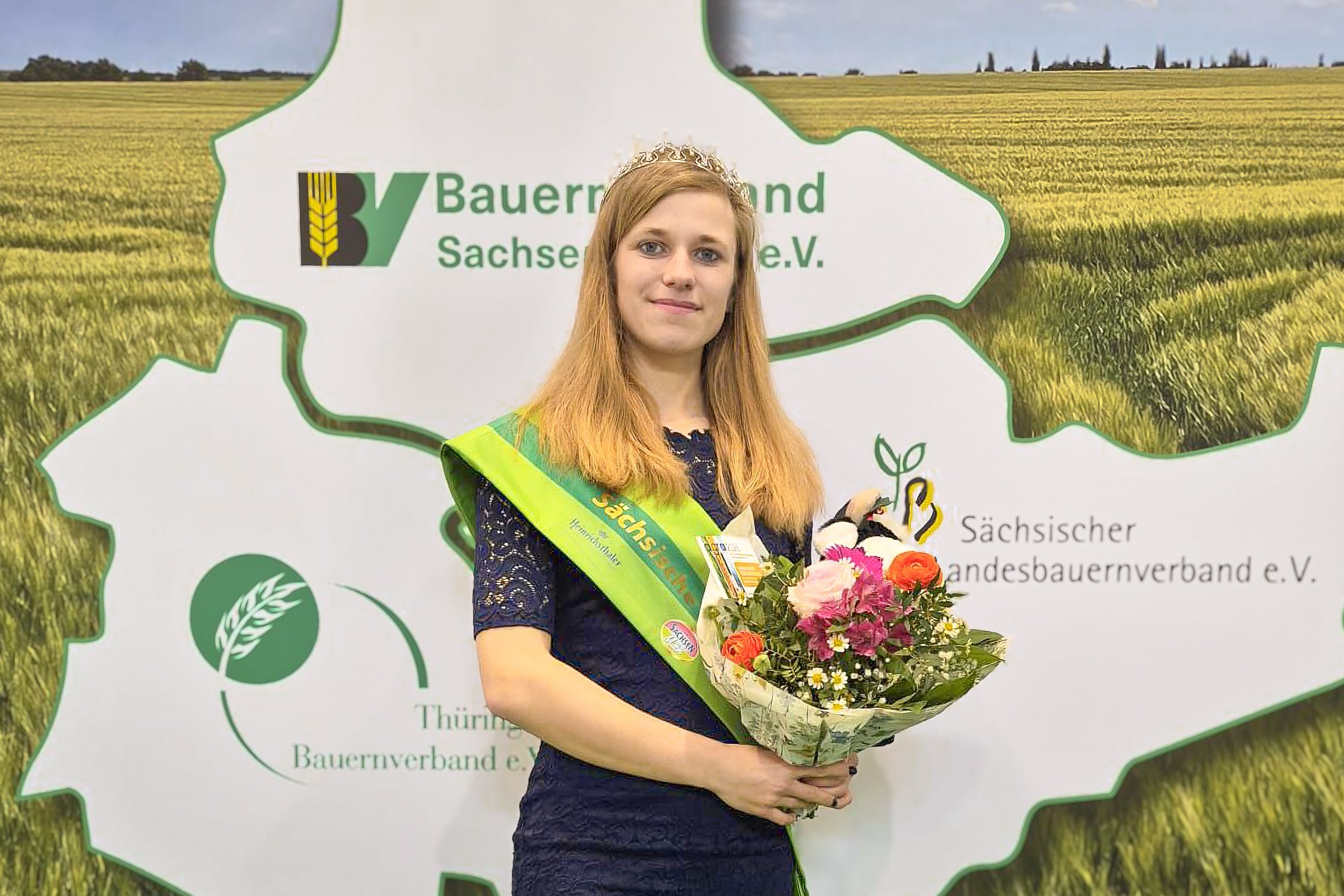 Eine Frau mit einer grünen Schärpe, einer Krone und einem Blumenstrauß in der Hand.
