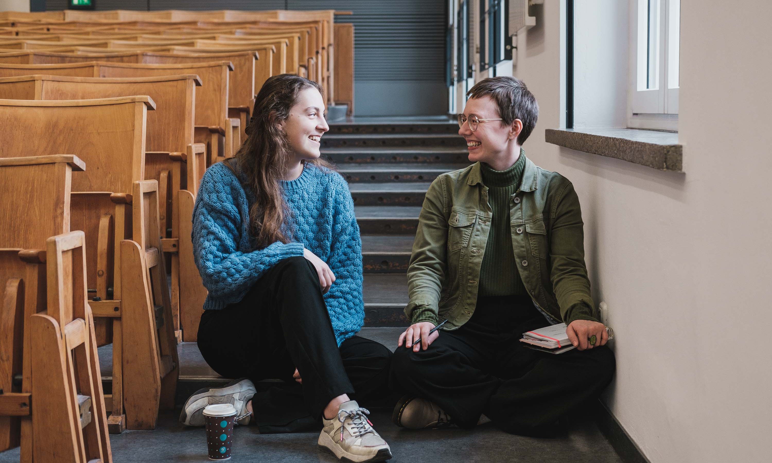 Zwei Frauen sitzen nebeneinander auf den Treppen eines Hörsaals und lächeln einander an.