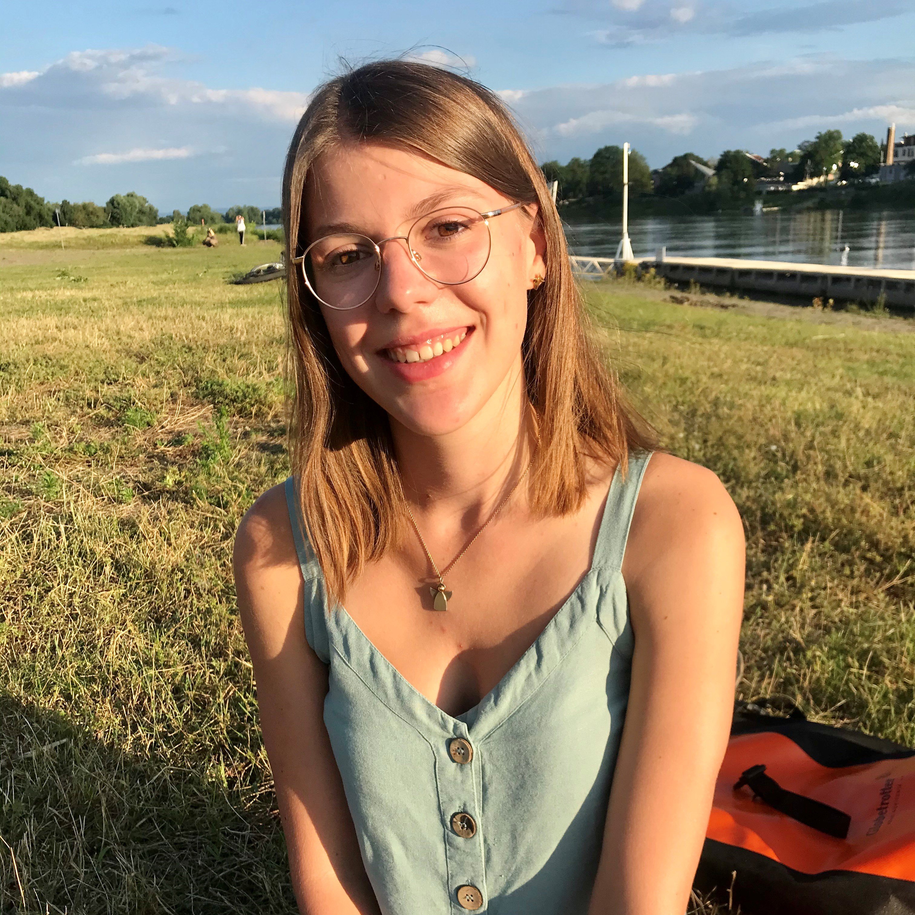 Foto, Studentin Sophie Cordes sitzt auf einer Wiese und lächelt. Im Hintergrund ist ein Fluss zu sehen. Sophie hat blonde, mittellange Haare, trägt eine Brille, hat Ohrringe und hat eine braune Augenfarbe. Sie trägt ein hellblaues Oberteil mit dünnen Trägern und Knöpfen. Zudem trägt sie noch eine Kette mit einem Anhänger.