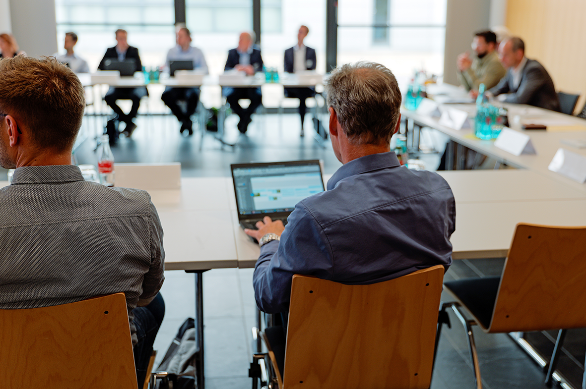 Menschen sitzen an einem U-förmigen Tisch und hören einem Vortrag zu. Im Vordergrund schaut ein Mann auf seinen Laptop