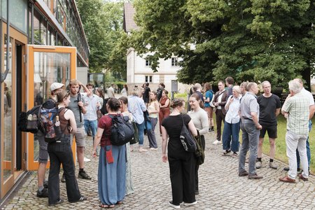 Menschen, die auf einem Platz vor einem Hörsaalgebäude stehen und sich unterhalten.
