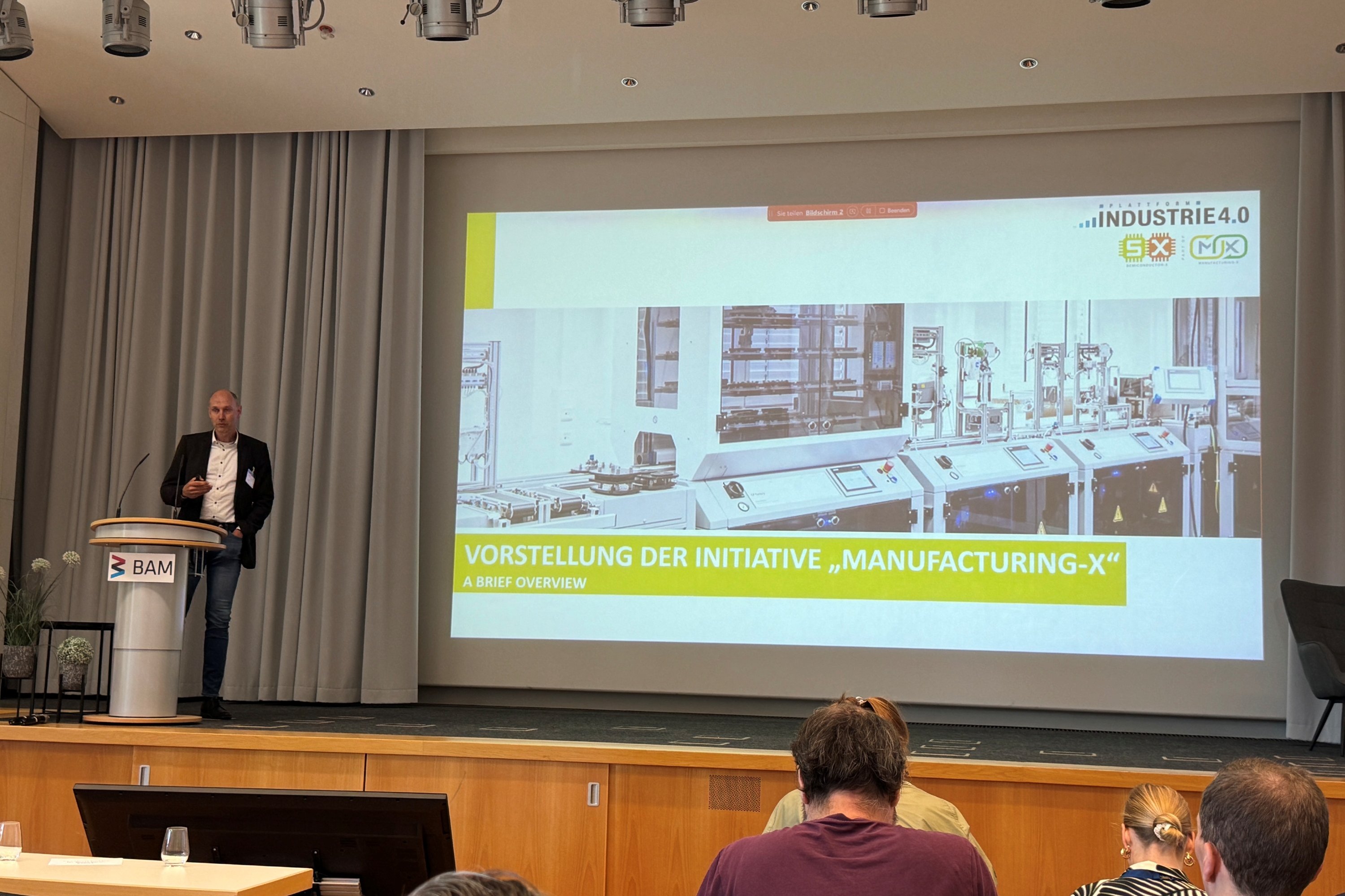 Prof. Dr. Dirk Reichelt steht an einem Pult auf einem Podium, im Hintergrund ist die Modellfabrik auf der Leinwand zu sehen