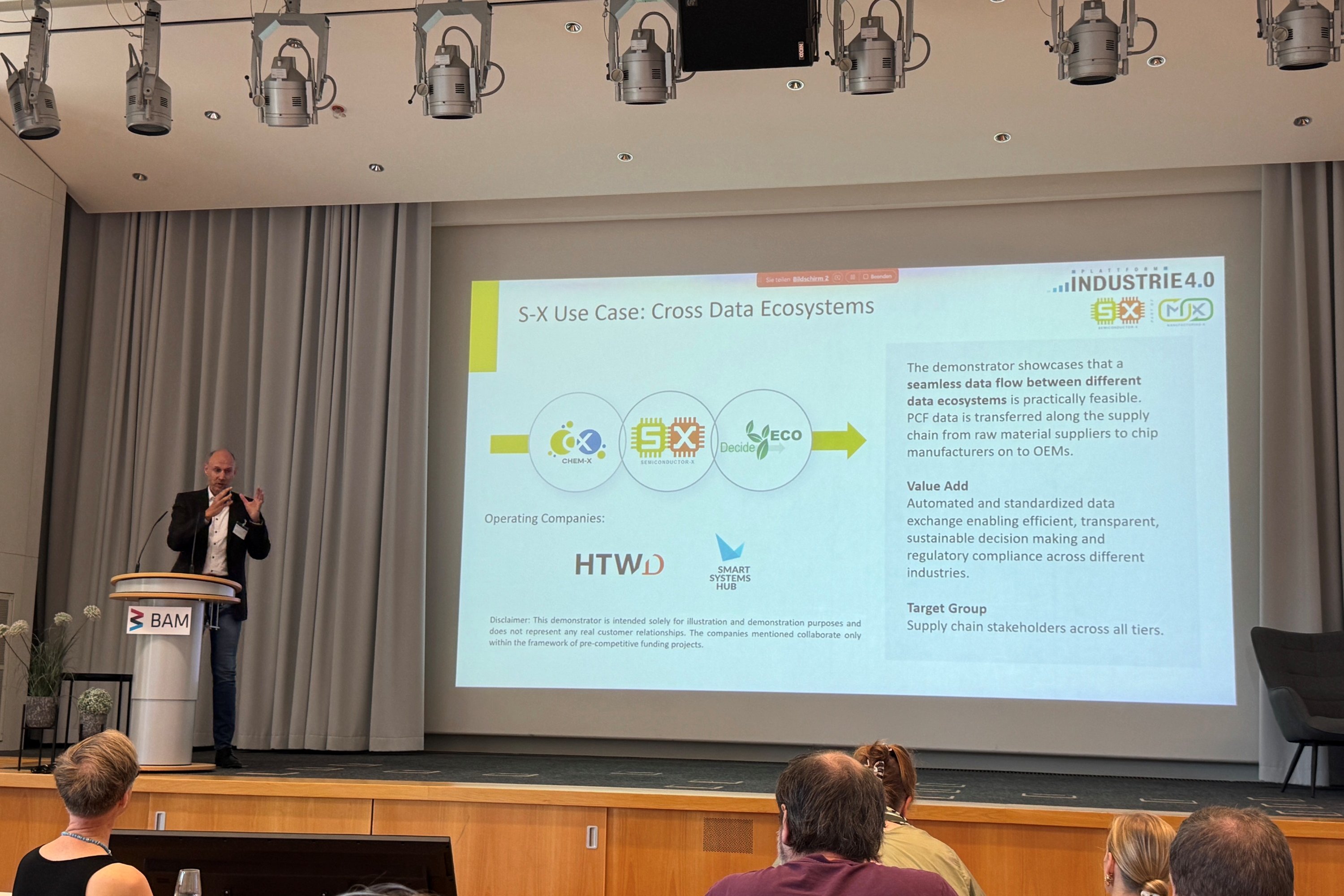 Prof. Dr. Dirk Reichelt steht an einem Pult auf einem Podium, im Hintergrund ist eine Folie der Präsentation zu sehen