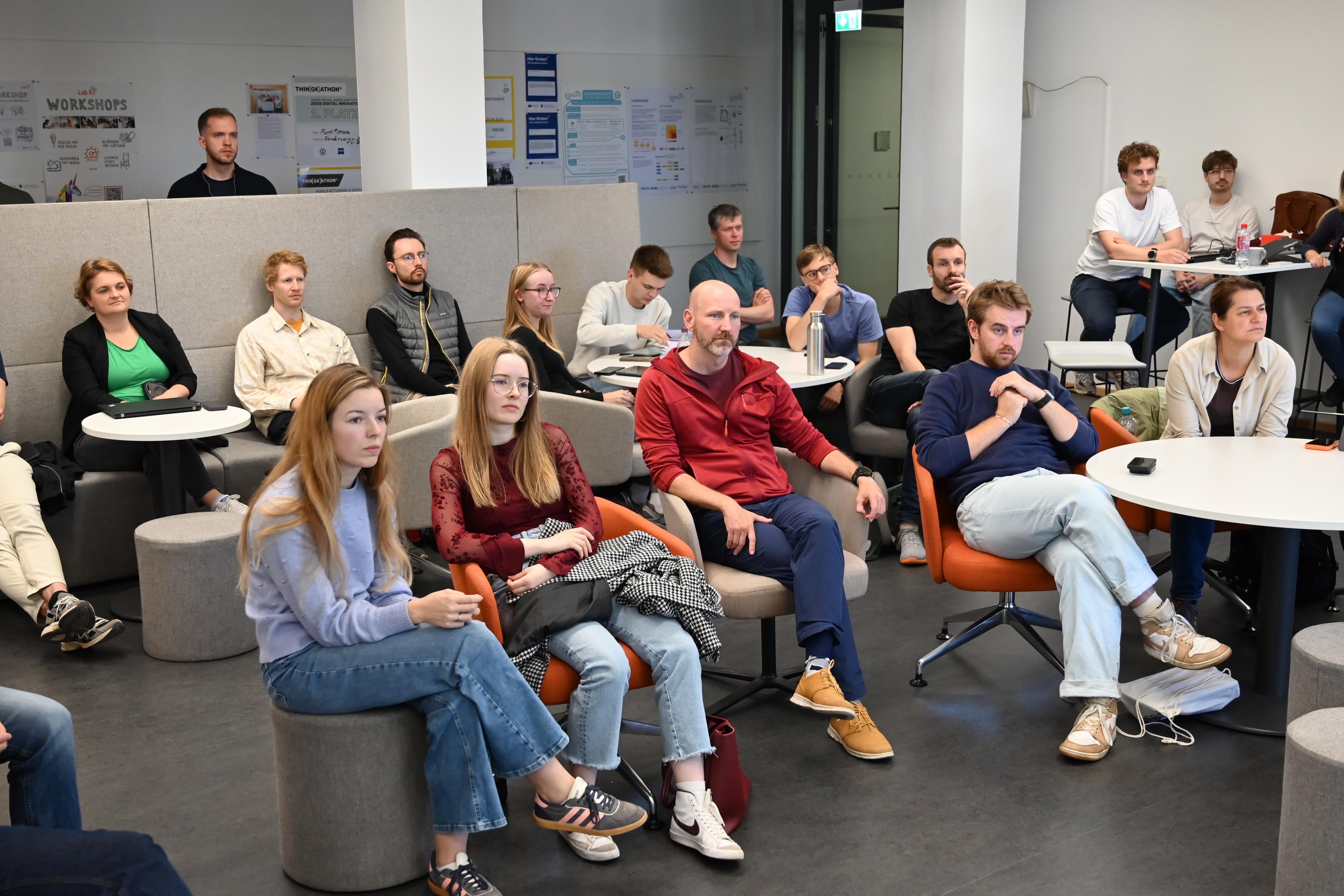 eine größere Gruppe interessiert schauender Menschen sitzt in einer Lounge und blickt in die selbe Richtung.