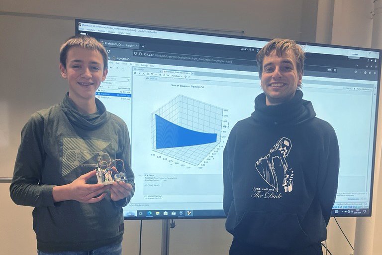 Ein Schüler und ein wissenschaftlicher Mitarbeiter stehen vor einem großen Monitor auf dddem Arbeitsergebnisse präsentiert werden