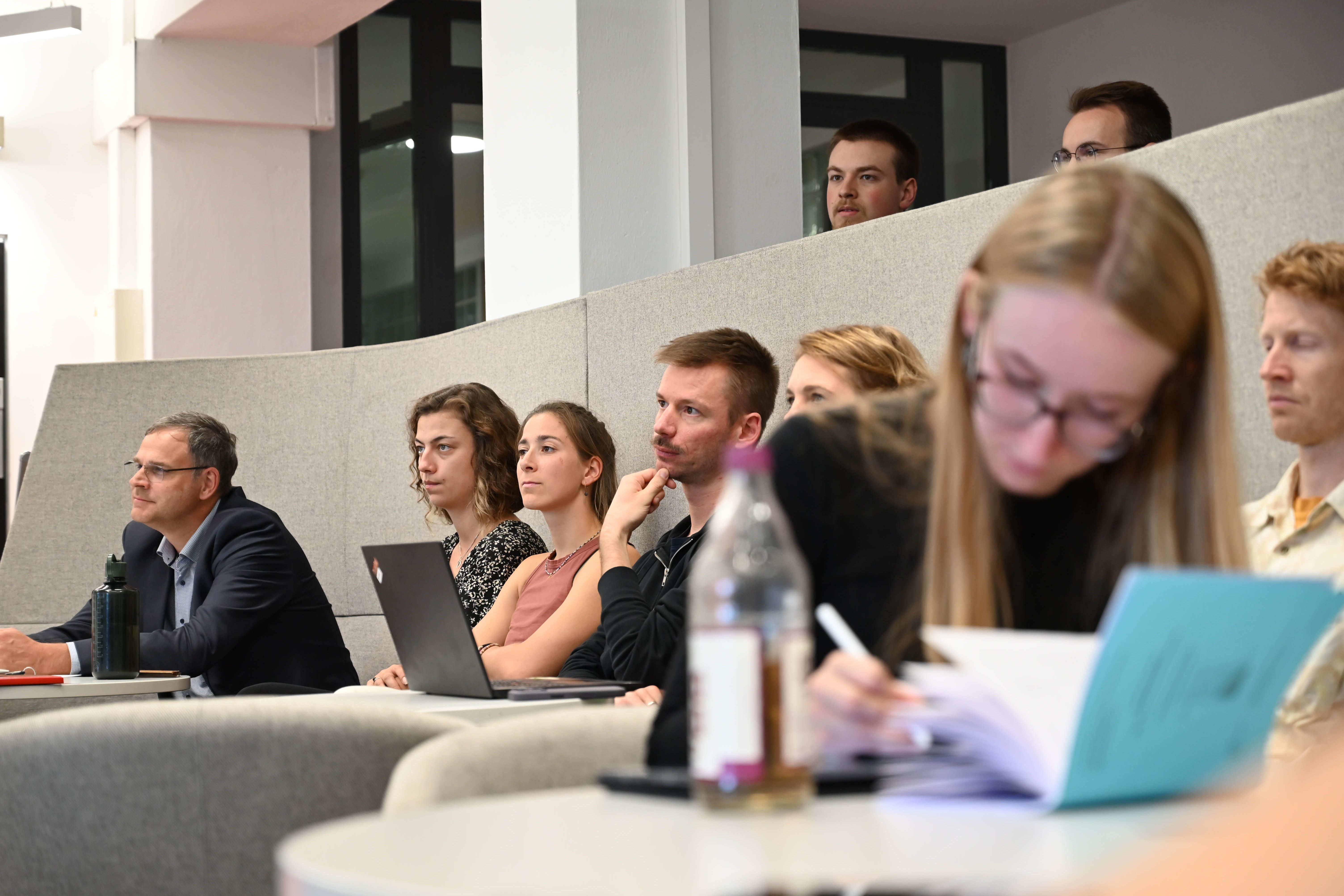 eine Gruppe interessiert schauender Menschen sitzt in einer Lounge und blickt in die selbe Richtung.