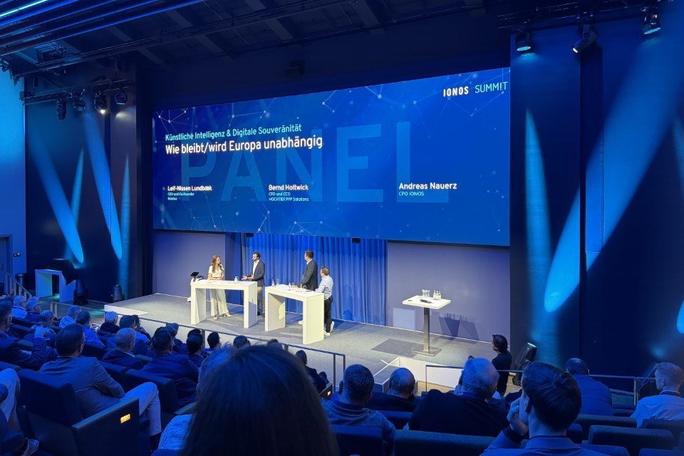 Es ist ein Podium zu sehen, auf dem sich mehrere Menschen unterhalten, im Vordergrund eine Menge Publikum